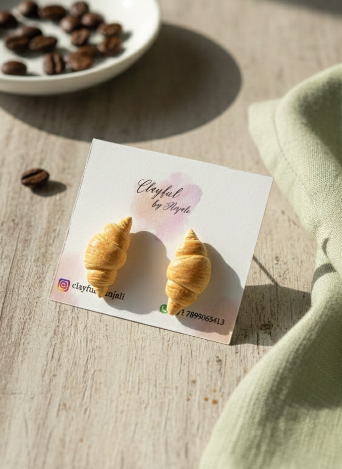 Hand-sculpted polymer clay stud earrings shaped like realistic golden-brown croissants with detailed flaky layers.