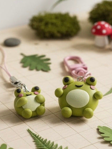 Two green frog-shaped keychains on a checkered surface with leaves and a mushroom in the background.
