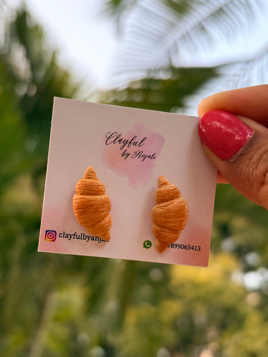 Hand-sculpted polymer clay stud earrings shaped like realistic golden-brown croissants with detailed flaky layers.